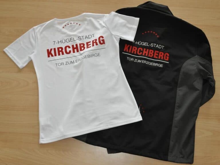 Auf dem Bild sind ein T-Shirt und eine Jacke zu sehen, die jeweils mit "7-Hügel-Stadt Kirchberg - Tor zum Erzgebirge" bedruckt sind