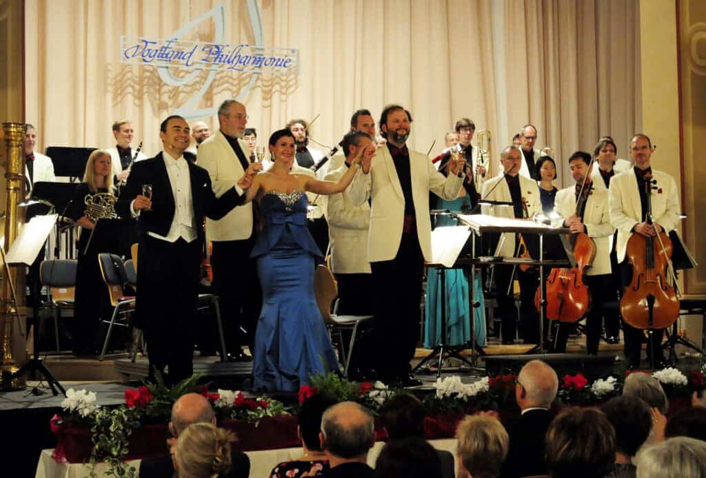 Auf dem Bild sind viele festlich gekleidete Menschen auf einer Bühne zu sehen. Einige von ihnen halten ein Musikinstrument in den Händen