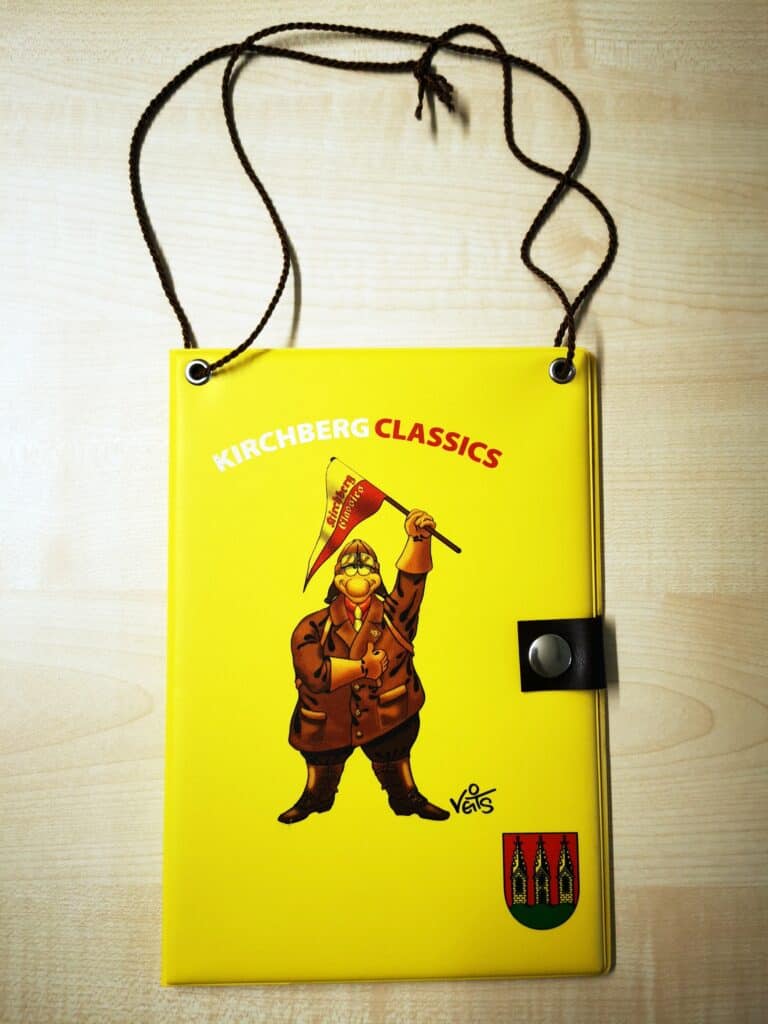 Auf dem Bild ist eine gelbe Fahrzeugmappe mit dem Schraubermichl, dem Kirchberg-Wappen und dem Schriftzug „Kirchberg Classics“ zu sehen
