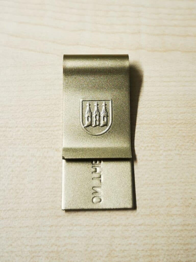 Auf dem Bild ist eine Geldklammer in Neusilberfarbe zu sehen. Auf der Vorderseite ist das Wappen der Stadt Kirchberg eingeprägt, auf der Rückseite der Schriftzug „On the road again“