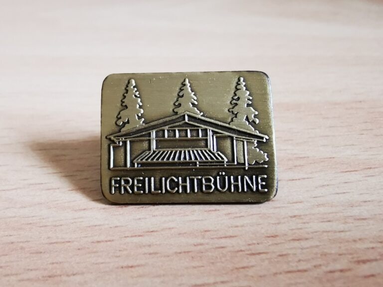 Auf dem Bild ist ein matter messingfarbener Pin mit der eingeprägten Freilichtbühne Kirchberg und dem Schriftzug „Freilichtbühne“ zu sehen
