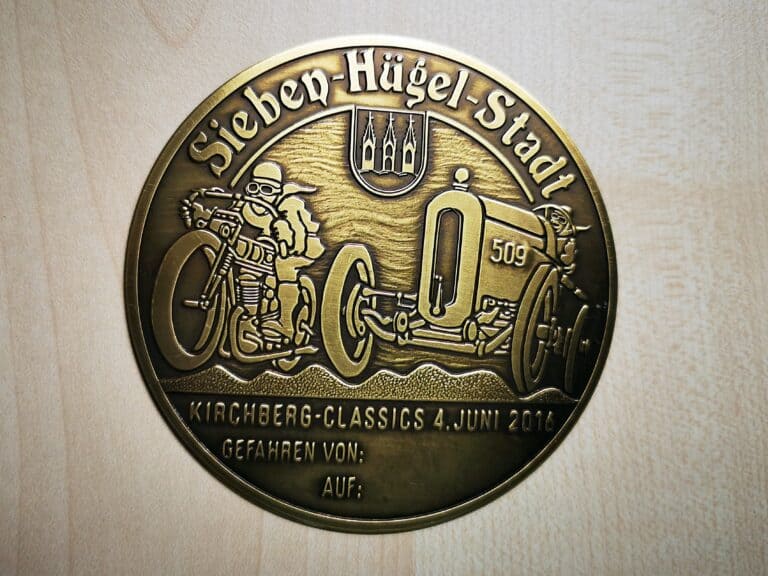 Auf dem Bild ist eine altgoldene Plakette „Kirchberg Classics“ 2016 mit zwei Oldtimern, dem Stadtwappen und mehreren Schriftzügen zu sehen