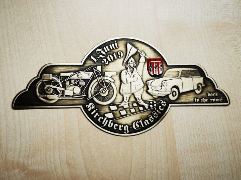 Auf dem Bild ist eine messingfarbene Plakette „Kirchberg Classics“ 2019 mit Oldtimern, dem Schraubermichl, Stadtwappen und eingeprägten Schriftzügen zu sehen