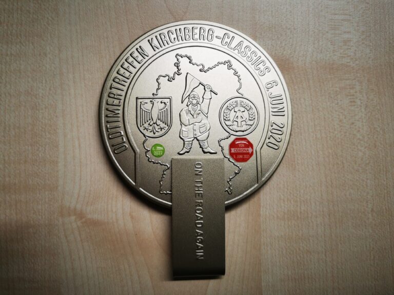 Auf dem Bild ist eine zweiteilige, silberne Plakette mit eingeprägten Motiven wie Deutschland-Umriss, Wappen und Datum sowie zwei Corona-bezogenen Aufklebern. Darauf liegt eine Geldklammer mit der Aufschrift „On the road again“.