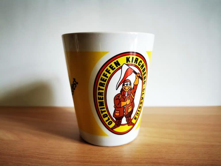 Auf dem Bild sind eine weiß-gelbe Tasse mit einer Illustration des Schrauber Michls und einer Aufschrift "Oldtimertreffen Kirchberg - Classics" zu sehen