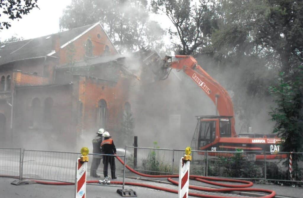 Auf dem Bild sind ein Bagger beim Abriss und Feuerwehrleute zu sehen, die einen Löschschlauch auf das eingerissene Gebäude richten