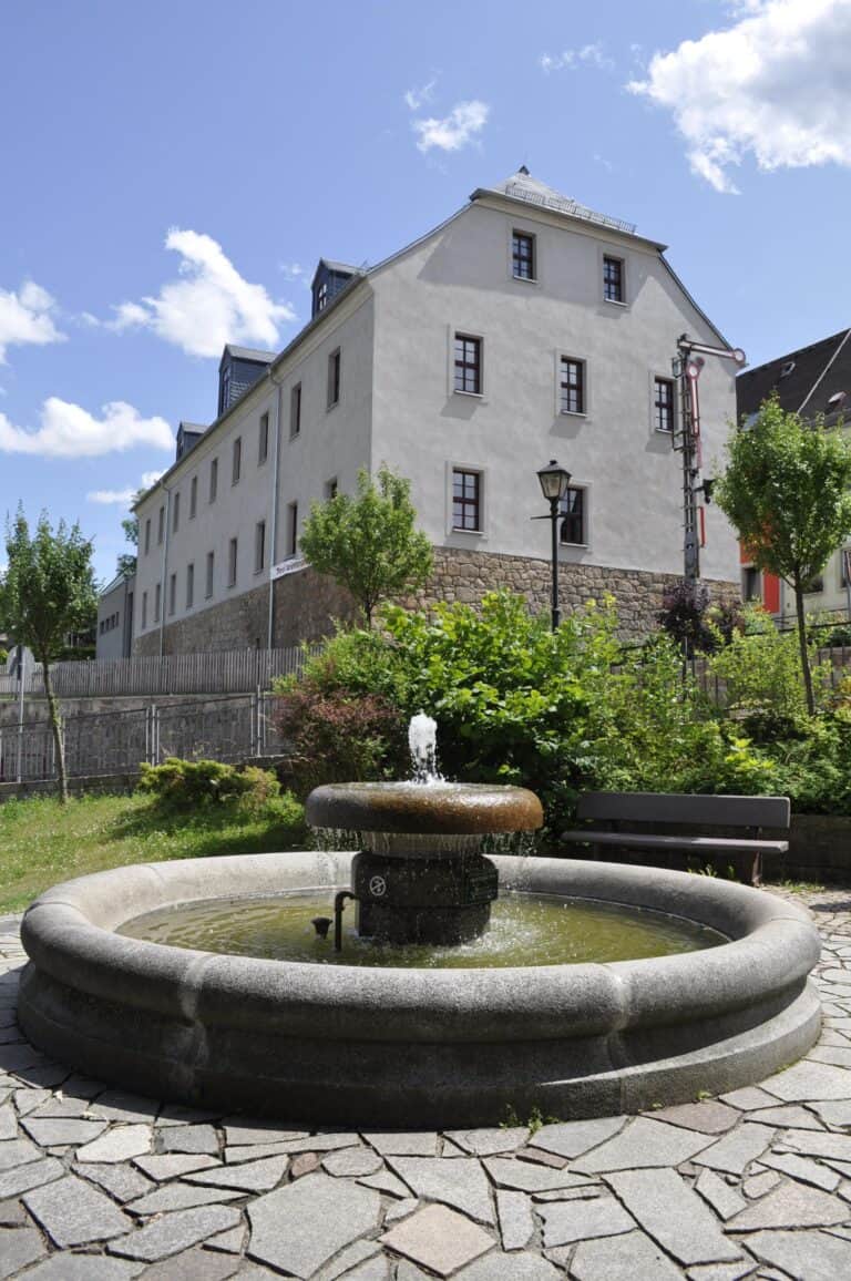 Auf dem Bild ist ein kleiner Springbrunnen im Vordergrund zu sehen. Dahinter erhebt sich das Meisterhaus