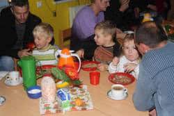 Auf dem Bild sind 3 Kinder zu sehen, die an einem Tisch essen und trinken. Im Hintergrund sind erwachsene Menschen zu sehen