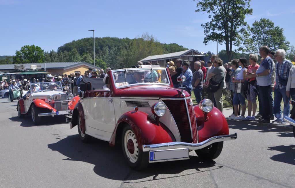 Auf dem Bild sind viele Zuschauer im Hintergrund zu erkennen, die zwei Oldtimer auf der Straße beobachten